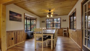 A wooden-paneled room with a dining table and chairs, a ceiling fan, and large windows with autumn foliage views.