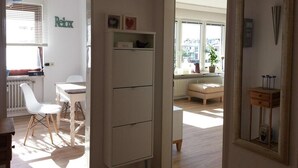 A modern, well-lit room with a white cabinet, a small table, and a chair. There is a window with blinds, a radiator, and a view of buildings outside. - Large roof terrace - Sun-drenched apartment - Elevator available (Bremerhaven)