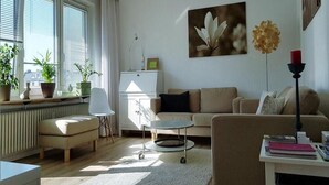 A living room with a sofa, a coffee table, a side table, a lamp, and a radiator. There are potted plants on the windowsill and a picture on the wall. - Large roof terrace - Sun-drenched apartment - Elevator available (Bremerhaven)