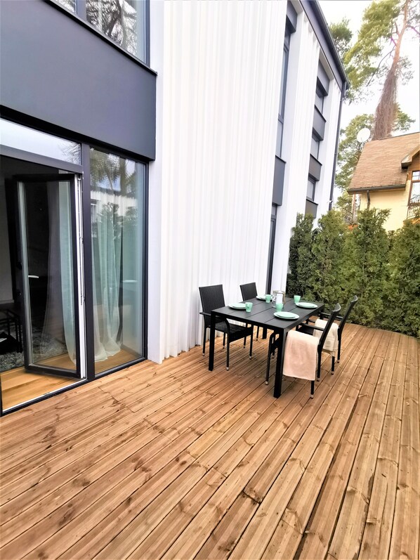 Een modern buiten eetgedeelte met een houten terras, een zwarte tafel met stoelen en een glazen deur naar een overdekt terras.