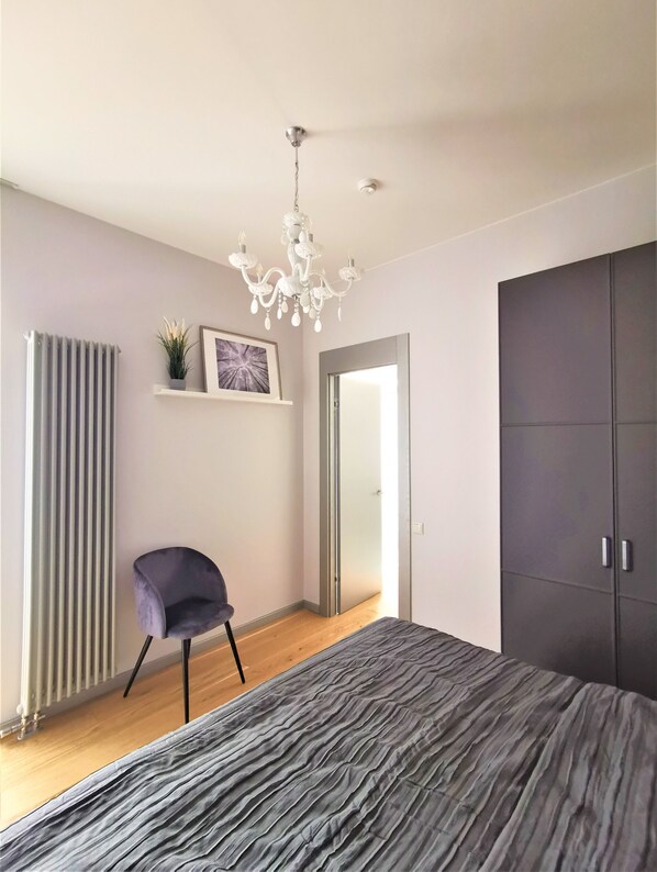 Een moderne slaapkamer met een groot bed, een stoel, een radiator, een plank met decoratie en een kledingkast.