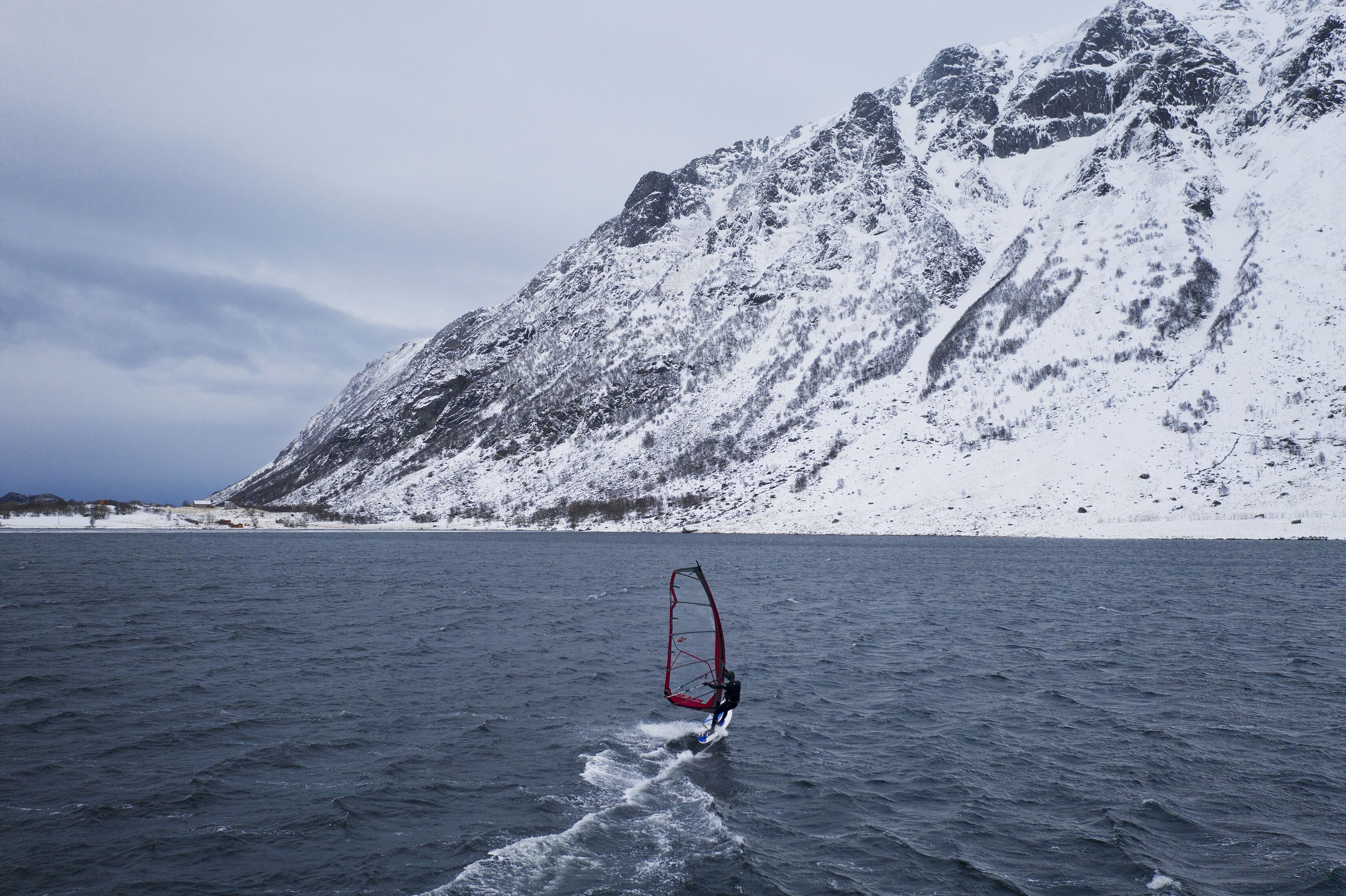 En person som åker vindsurfing på en sjö eller ett hav med ett snötäckt berg i bakgrunden.