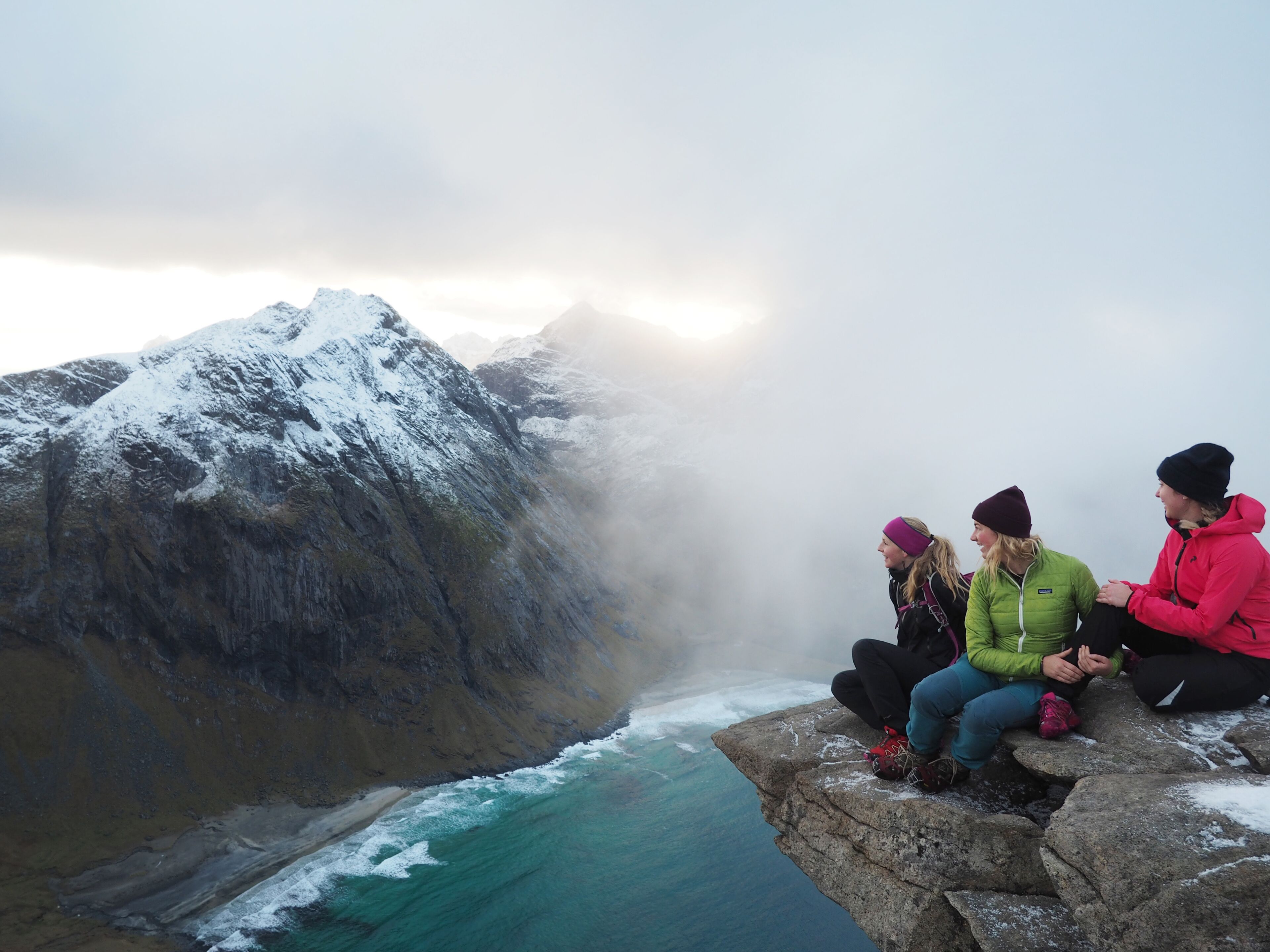 Tres personas sentadas en un saliente rocoso con vistas a una zona costera y montañas al fondo.