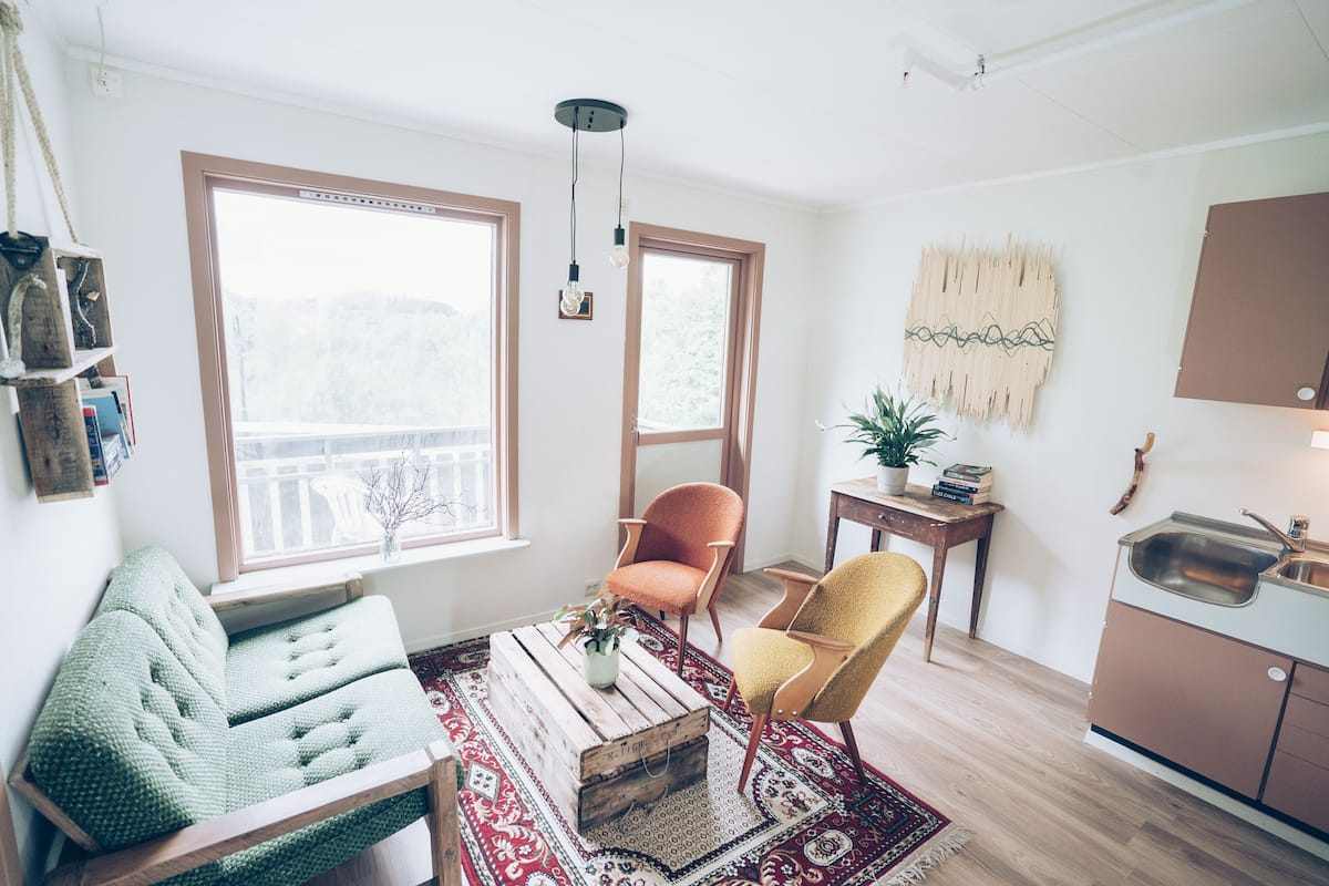 A cozy living room with a green sofa, a wooden coffee table, and a yellow armchair. There is a large window with a view of trees, a small table with a plant, and a kitchen area with a sink and cabinets.