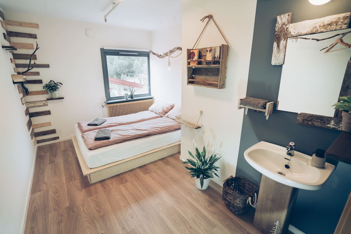 A bedroom with a bed, a window, a bedside plant, a wooden shelf, and a bathroom with a sink.