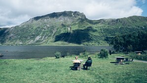 Vista al lago con una montaña al fondo, una mesa con sillas y una persona caminando a lo lejos.