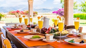 Disposição de mesa para o café da manhã para quatro pessoas, com pratos, xícaras e copos de suco de laranja, e uma vista panorâmica ao fundo.