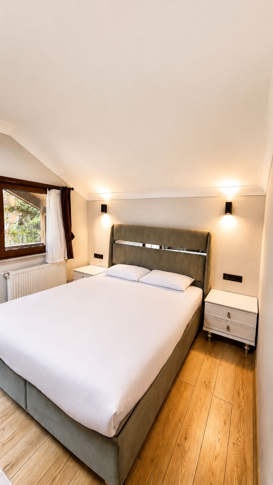 Une chambre à coucher avec un grand lit, des tables de chevet, une fenêtre avec des rideaux et des lampes fixées au mur.