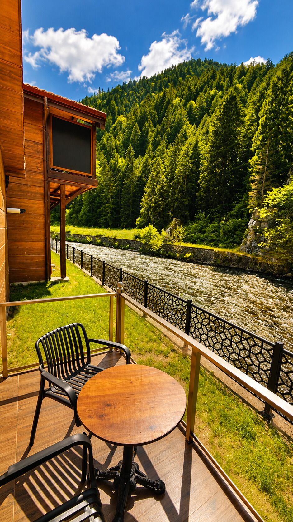 Un terrazzo in legno con tavolo e sedie affacciato su un fiume, con una collina boscosa sullo sfondo.
