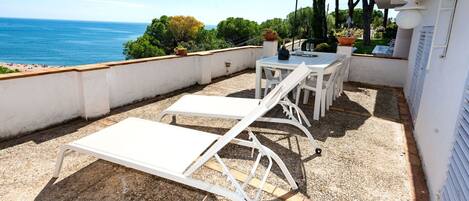 An outdoor patio with white lounge chairs and a table, overlooking the sea.