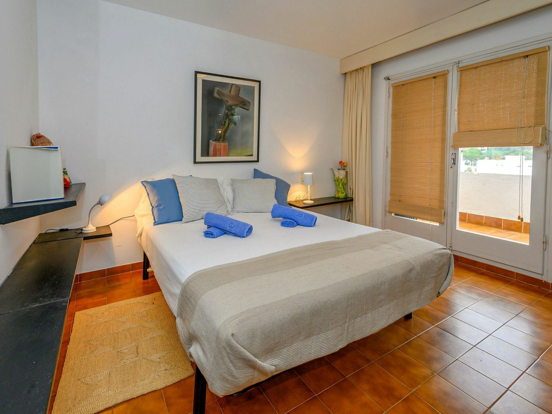 A hotel room with a large bed, a desk, a chair, a window with blinds, and a framed picture on the wall.