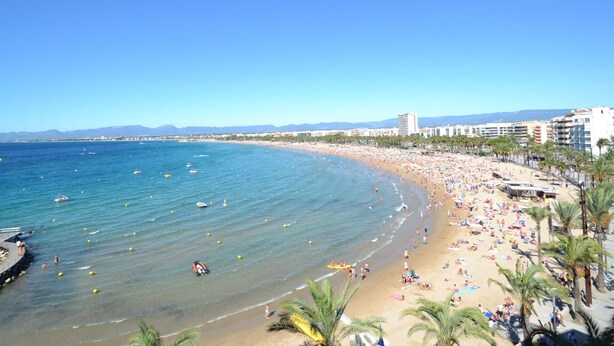 Une plage avec une eau d’un bleu limpide, des palmiers et des bâtiments en arrière-plan.