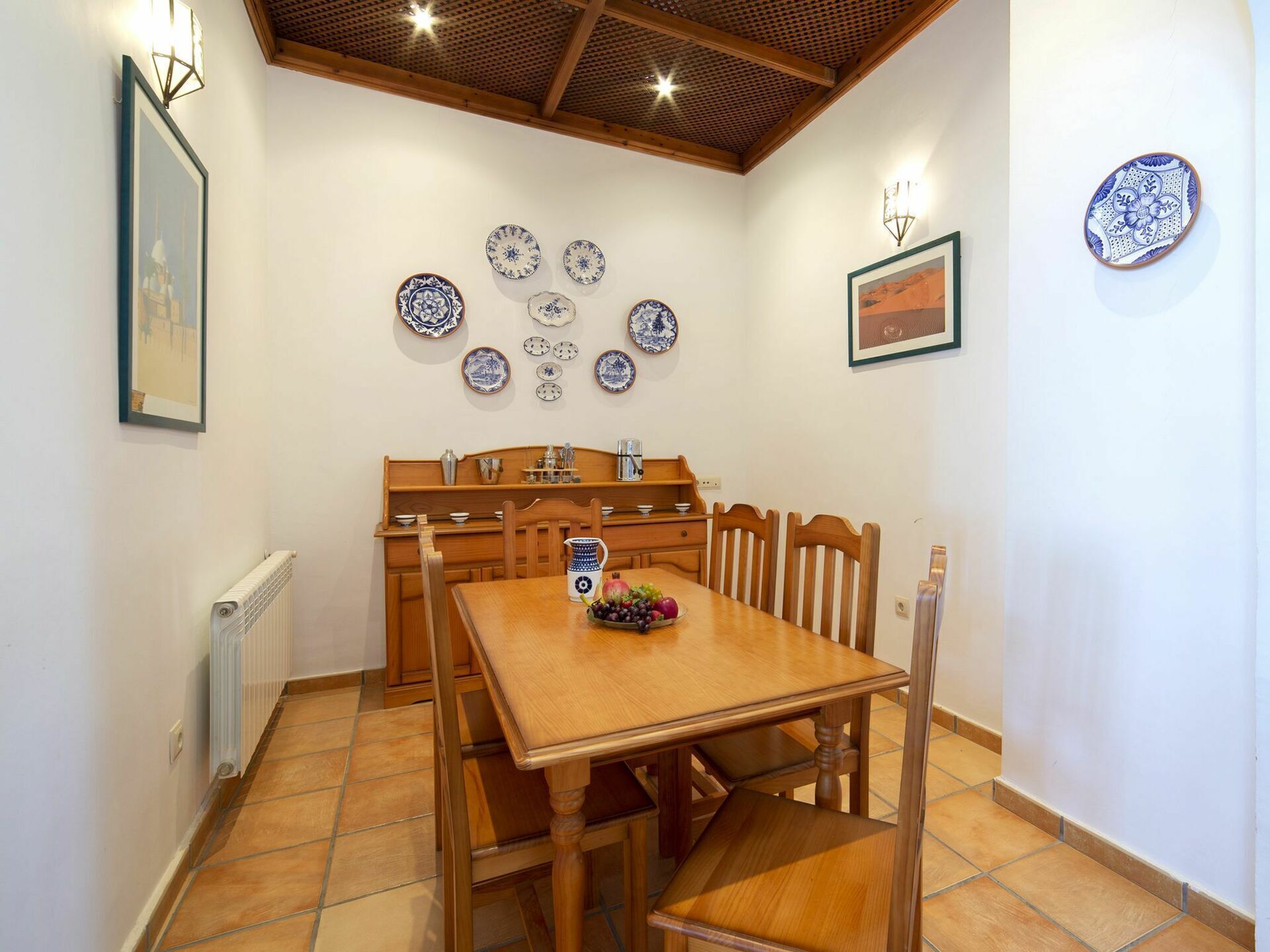 A dining area with a wooden table and chairs, a tiled floor, and a wall decorated with plates and framed pictures.