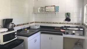 Une cuisine compacte avec des meubles blancs, un four à micro-ondes, une machine à café et une cuisinière à gaz. Des étagères au-dessus du comptoir servent à ranger des bocaux, et un panneau est accroché au mur.