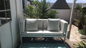 Un espace extérieur aménagé avec des sièges blancs, agrémenté d’un tapis à motifs, entouré de plantes en pot et d’une clôture en bois.