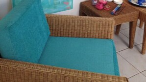 Une pièce comprenant un canapé de couleur turquoise, un fauteuil en osier, un tableau représentant un flamant rose, une lampe et une petite table sur laquelle se trouvent divers objets.