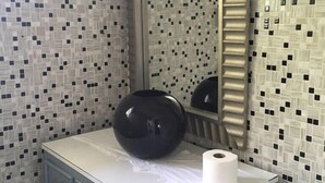 Uma casa de banho com parede revestida em azulejos, espelho, lavatório com um vaso e rolo de papel higiénico.