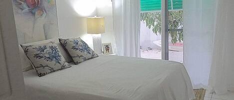 Un dormitorio con cama, mesita de noche, lámpara, un cuadro y ventana con cortinas.