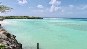 Paisagem costeira com águas turquesa cristalinas, uma praia arenosa e um afloramento rochoso com vegetação rasteira.