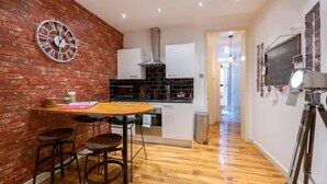 Una cocina compacta con pared de ladrillo, un reloj redondo, una mesa de madera con taburetes altos y un pequeño cubo de basura.