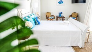 Un dormitorio ordenado con una cama, dos sillas, una mesita y un televisor en la pared.