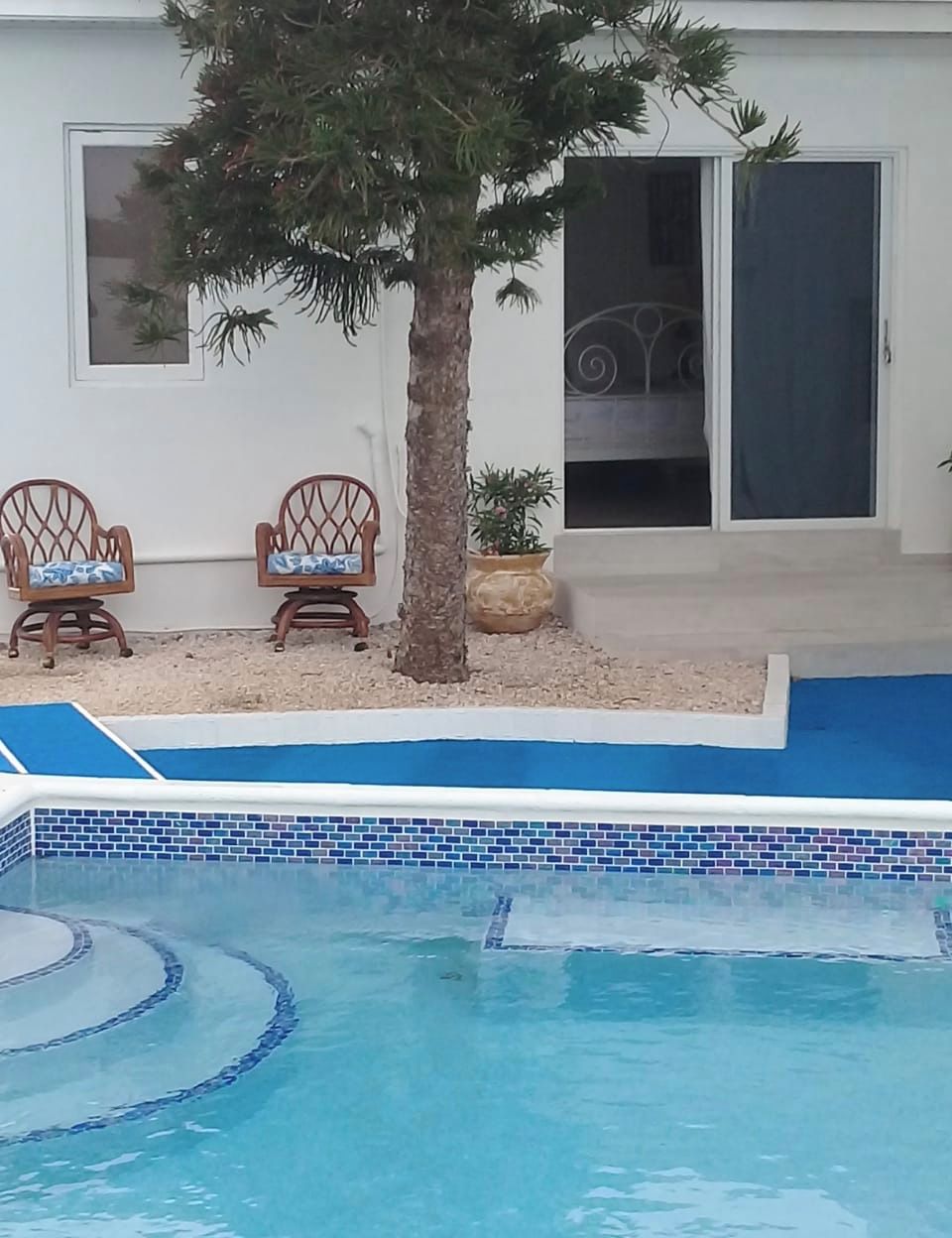 Una piscina amb una vores enrajolada, dues cadires de fusta amb coixins, una palmera i un edifici amb una porta corredissa de vidre.