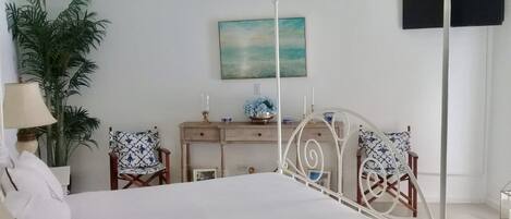 A bedroom with a bed, a desk, chairs, a plant, and artwork on the wall.