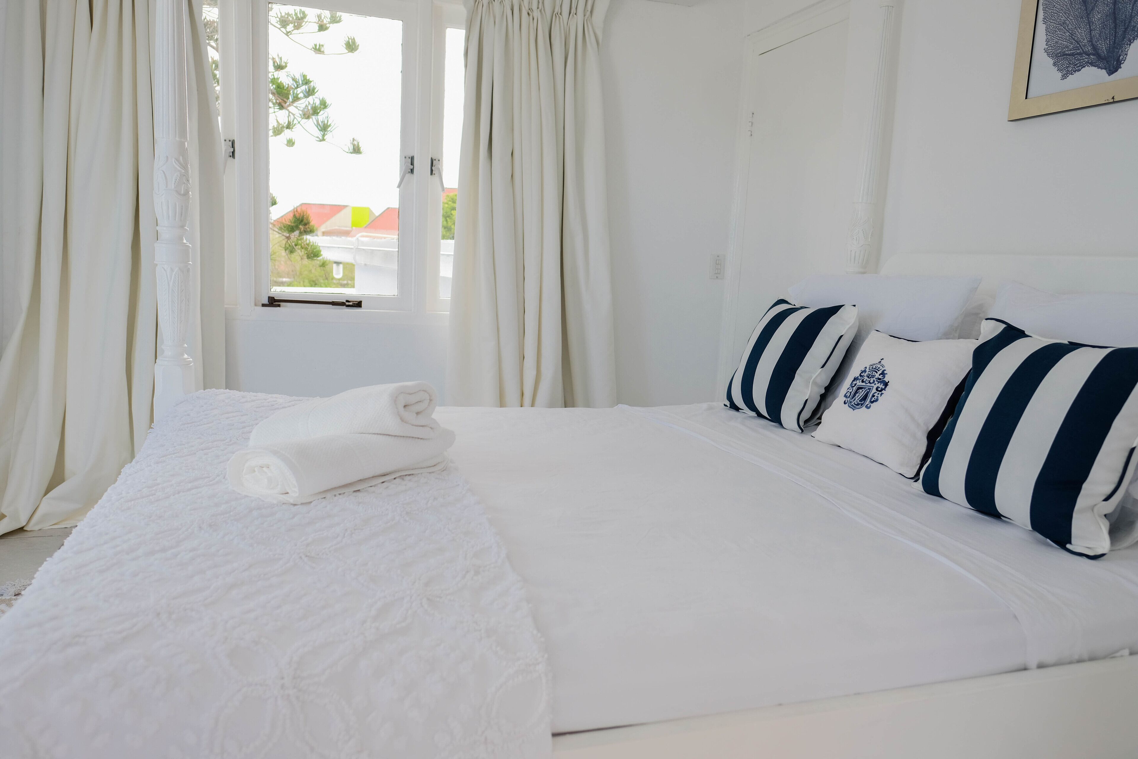 A neatly made bed with white linens and striped pillows, a window with curtains, and a framed picture on the wall.