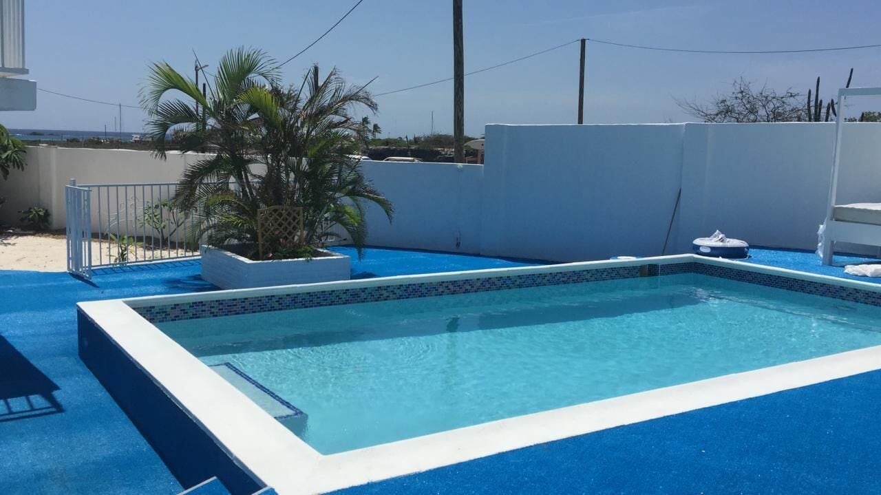 Una piscina rectangular con borde de azulejos azules, rodeada por un muro blanco y un suelo azul.