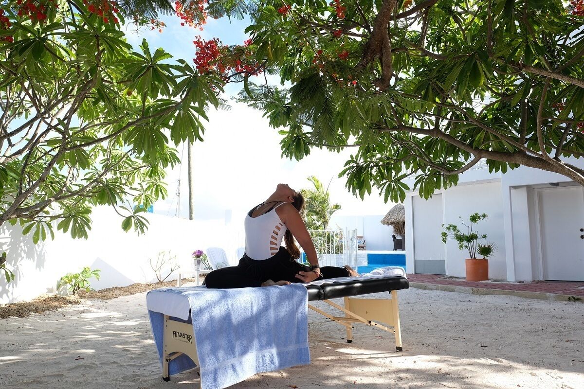 Eine Person führt eine Yoga-Pose auf einer Massageliege im Freien aus, im Hintergrund ist ein Schwimmbad und ein Gebäude zu sehen.