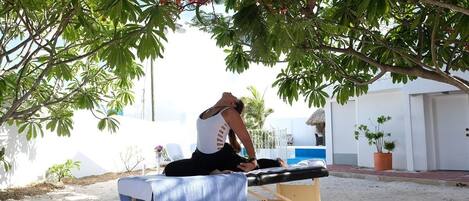 Eine Person führt eine Yoga-Pose auf einer Massageliege im Freien aus, im Hintergrund ist ein Schwimmbad und ein Gebäude zu sehen.