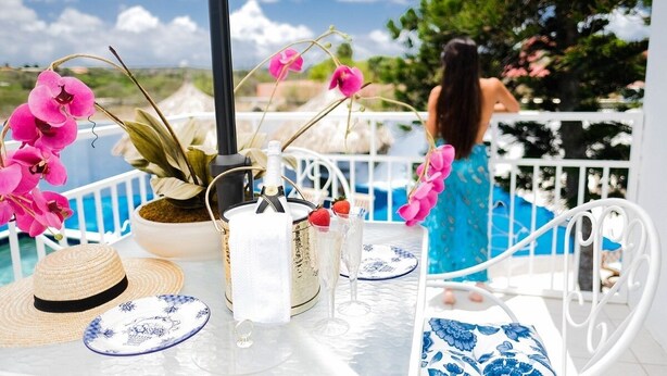 A table set with a bottle of champagne, a glass, a straw, a plate, and a hat, overlooking a pool and greenery. - Ocean Front Property - Villa 5 Aruba Stunning (Savaneta)