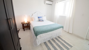 Een slaapkamer met een bed, nachtkastje, lamp, raam met gordijnen en een airconditioning.