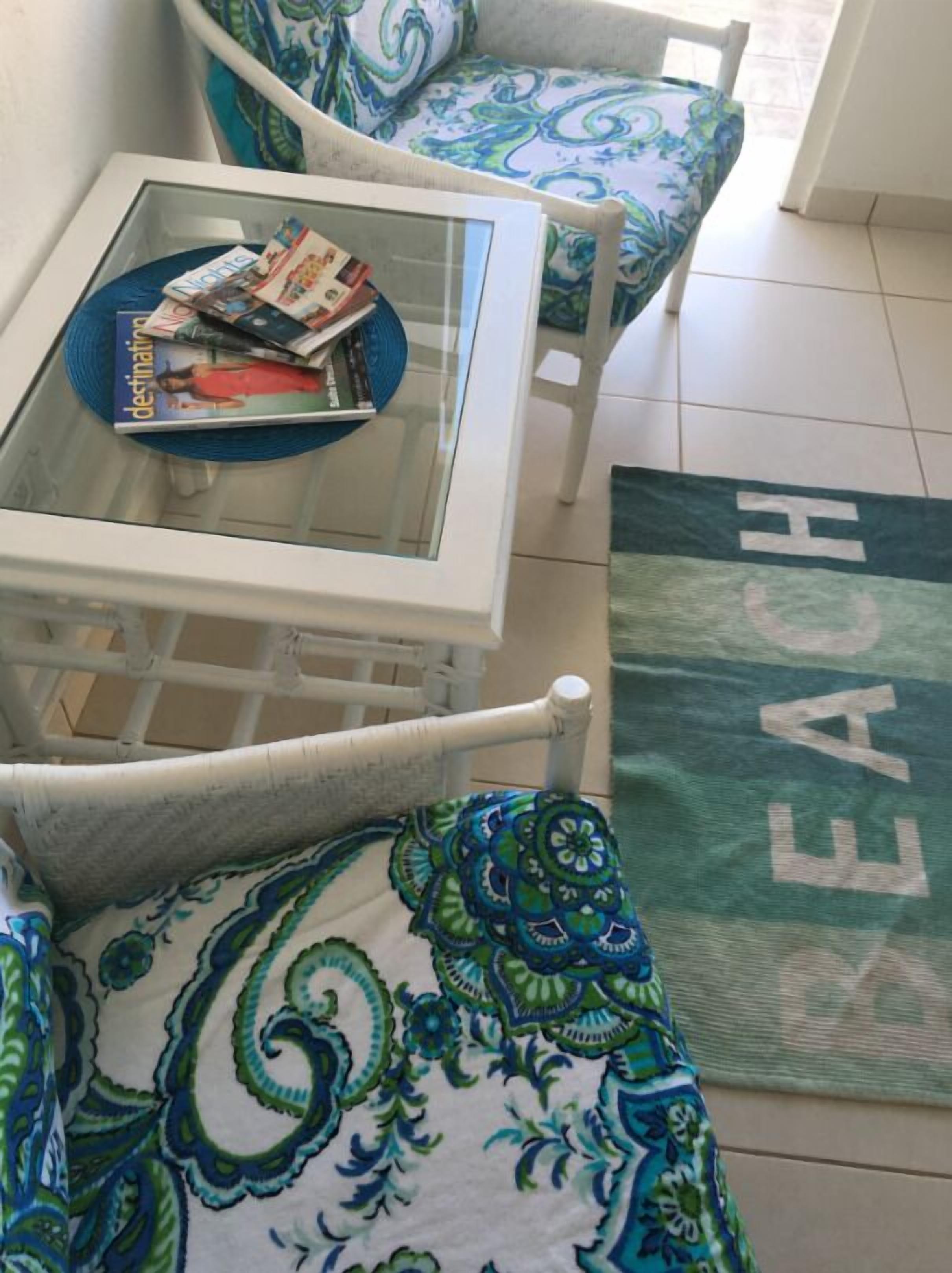 A room with a glass-top table holding magazines, a patterned rug on the floor, and a white chair with a blue and green design.