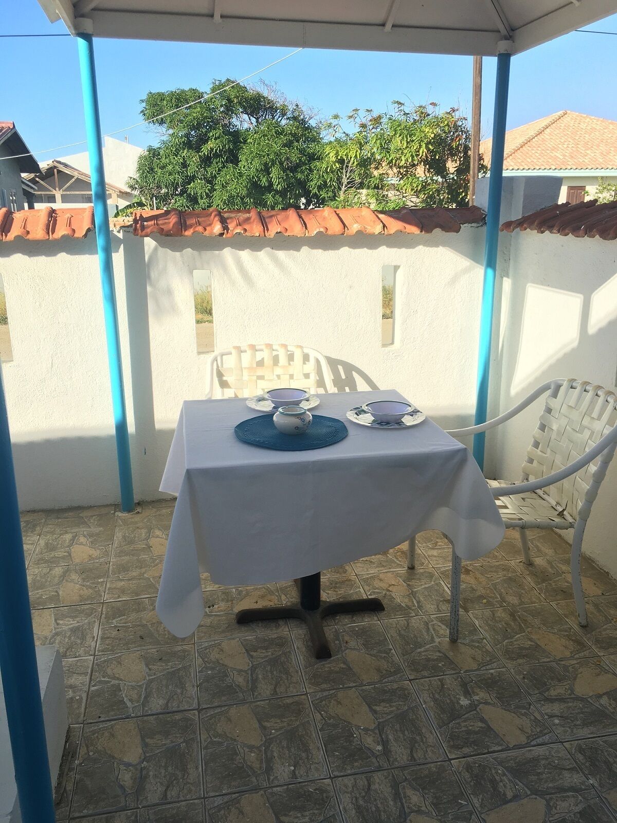 Un patio couvert avec une table dressée pour deux personnes, agrémenté d’une nappe blanche et de sets de table bleus.