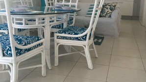 Uma área de refeições com mesa de vidro e cadeiras brancas com almofadas azuis, um sofá cinza com almofadas estampadas e uma cozinha com armários brancos e painel de azulejos.