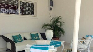 Une pièce aux murs blancs, avec un canapé blanc, deux fauteuils blancs, une plante en pot et une fenêtre à motifs.
