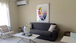 Un salon moderne avec un canapé gris, une table basse blanche et un fauteuil à bascule blanc. Un tableau abstrait aux couleurs vives est accroché au mur, et un panneau est posé sur le sol.