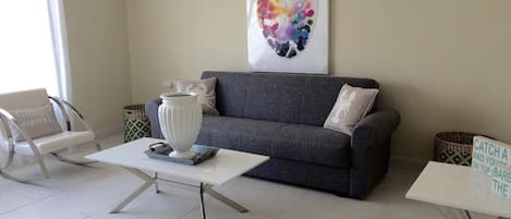 A modern living room with a grey sofa, white coffee table, and a white rocking chair. There is a colorful abstract painting on the wall and a sign on the floor.