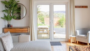 Un salon comprenant un canapé, une table basse avec un jeu d’échecs, un meuble en bois, un miroir et une porte-fenêtre coulissante donnant sur une terrasse extérieure avec une table et des chaises.