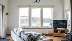 Un salon comprenant un canapé, un téléviseur, une cheminée et de grandes fenêtres offrant une vue sur des collines et l’eau.