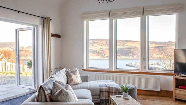 Un salon moderne avec un canapé, une table basse et un téléviseur. De grandes fenêtres offrent une vue sur un lac et des collines.