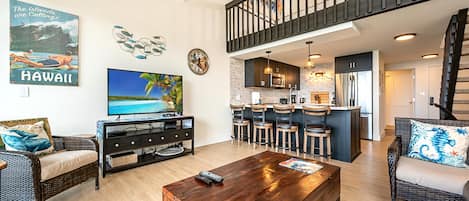 A spacious living area with a TV, wooden coffee table, wicker furniture, a kitchen with bar stools, and a staircase.