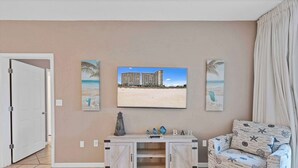 A living room with a beige wall, a flat-screen TV mounted above a wooden console table, a beach-themed sofa, and a door leading to another room.