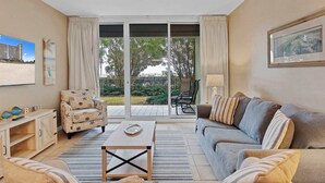 A living room with a sofa, armchair, coffee table, and a view through a sliding glass door to a patio area.
