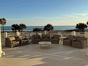 Relax in this beachside seating area with a round table and wicker furniture, offering stunning views of the ocean and palm trees.
