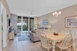 Relax in this open-plan living space with a dining table, sofa, and ceiling fan, offering a view of lush greenery through the sliding glass door.