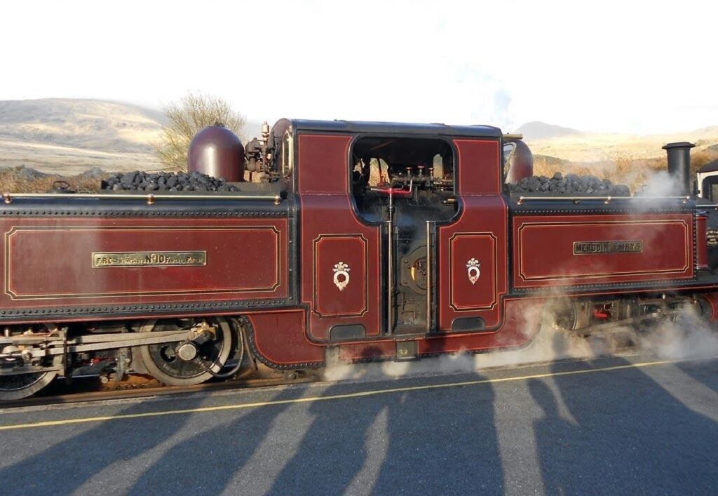 Eine Dampflokomotive mit der Aufschrift "HERTFORD EMPIRE" auf der Seite, fährt auf Gleisen durch eine bergige Landschaft.