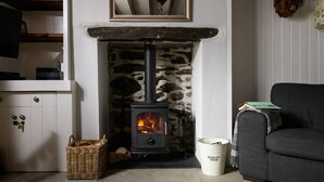 A cozy living room with a stone fireplace, a grey sofa, a wicker basket, and a bucket labeled "Kindling Wood".