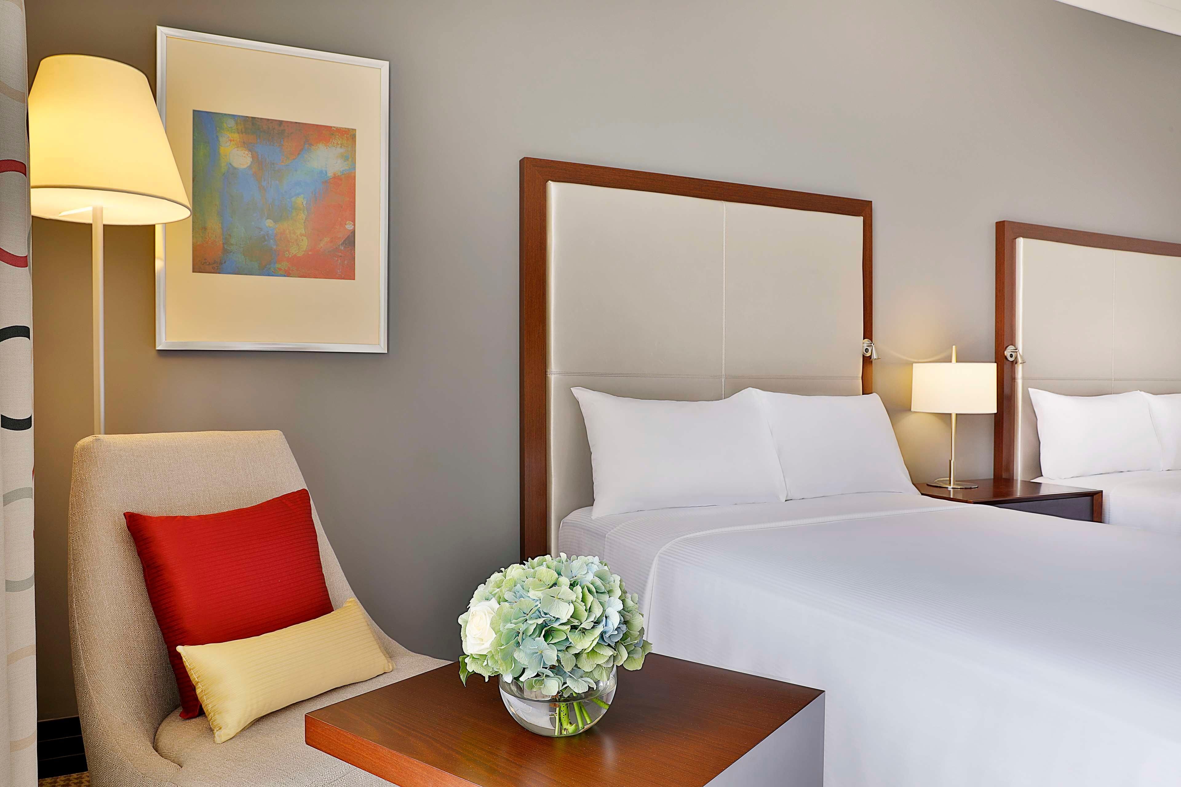 A hotel room with a bed, a chair with a red pillow, a bedside table with a vase of flowers, a lamp, and a framed abstract painting on the wall.